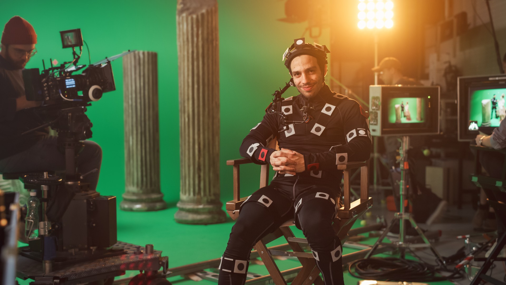 Handsome Smiling Actor Wearing Motion Capture Suit and Head Rig having Lunch Break, Sitting on Chair, Looking at Camera. Studio High Budget Movie. On Film Studio Period Costume Drama Film Set